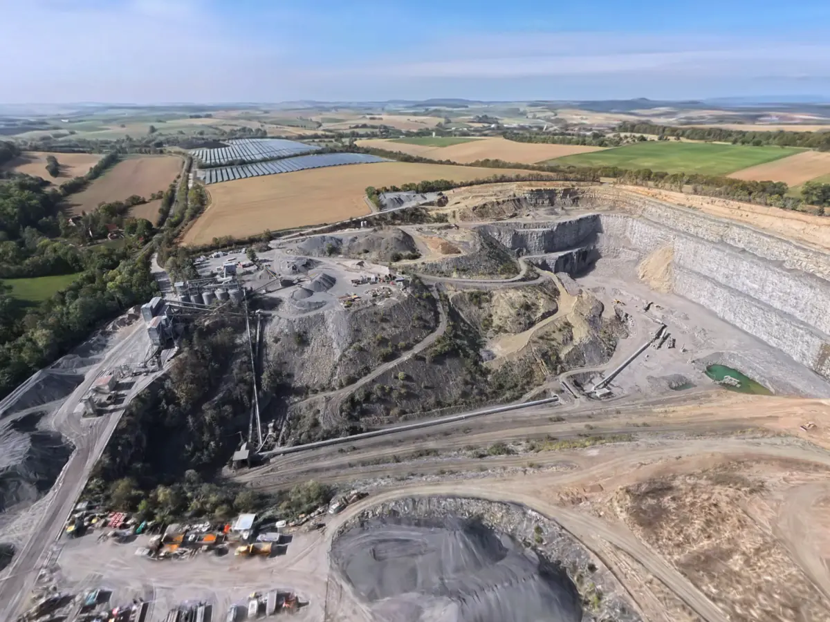Oblique aerial view of the quarry from high up — on the right the tall working face of pale grey, horizontally bedded limestone, stepped down in several benches, with a yellowish layer along the top; on the floor a small green pond with a floating platform and a machine from which a long conveyor belt leads out of the pit. On the left the processing plant with a row of round silos, steel frames and conveyors, next to it conical grey stockpiles of crushed stone and a yard full of machinery; in the middle a dark grey, sparsely overgrown spoil slope with a zigzag track. All around harvested fields, hedgerows and a wooded valley, beyond them a solar park and rolling country under a slightly hazy sky.