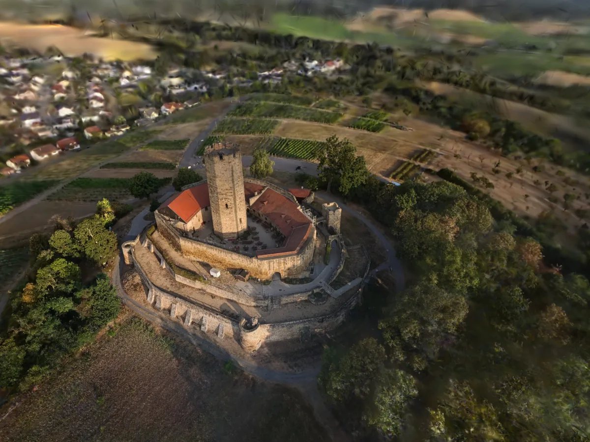Schräge Luftaufnahme der ganzen Burganlage — in der Mitte der achteckige Bergfried aus hellem Buckelquaderstein, um den Innenhof rot gedeckte Ringbauten, darum die Wehrmauer mit Strebepfeilern und ein umlaufender Wallgraben, dahinter Weinberge, Streuobst und der Ort Weiler